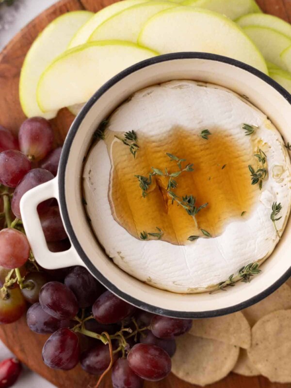 baked brie wheel in a small round pot with thyme and honey next to crackers and fruit on a wooden board.